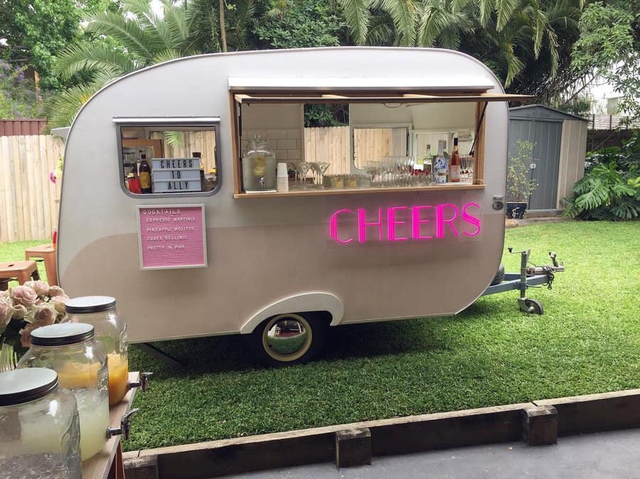 Retro caravan bar on grass with a "CHEERS" neon sign, offering drinks surrounded by greenery and wooden fence in the background