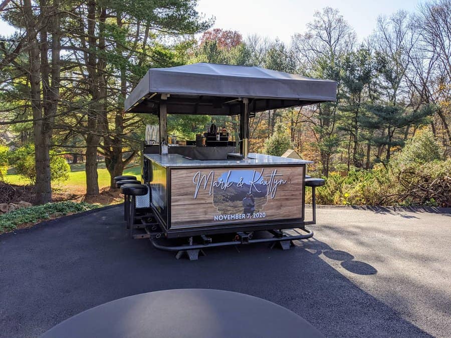 Mobile bar cart with stools on a paved area, surrounded by trees, personalized with names and a date