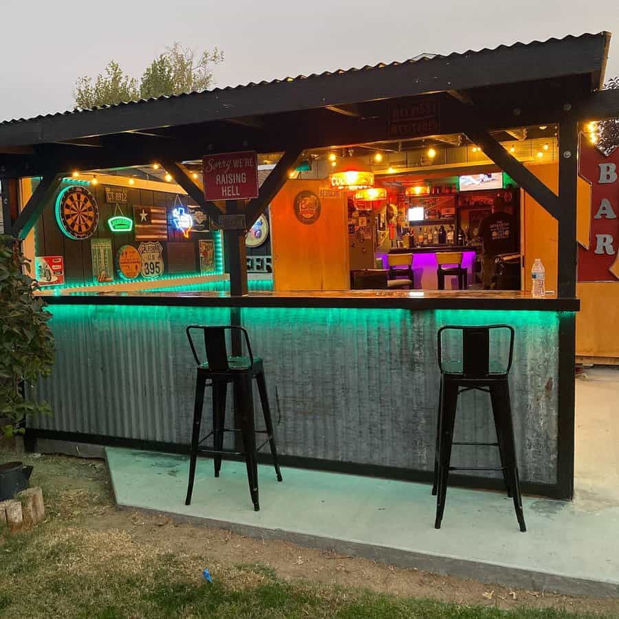 Outdoor bar with metal structure, two chairs, neon lights, and dartboard; interior decorated with colorful signs and memorabilia