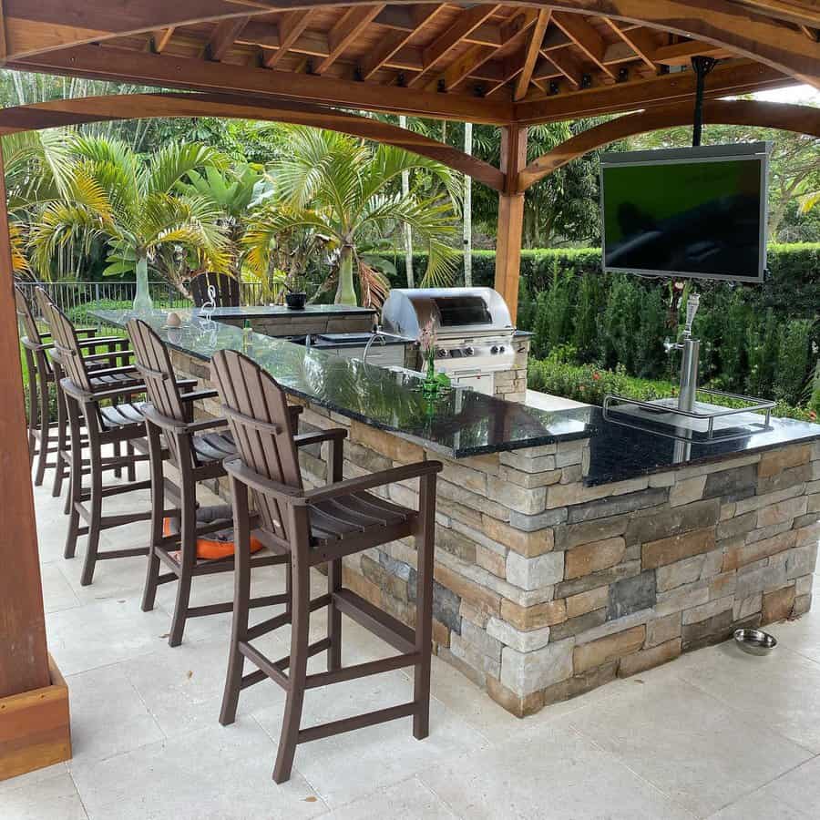 Outdoor kitchen with stone bar, four wooden bar stools, grill, and wall-mounted TV under a wooden pergola surrounded by greenery