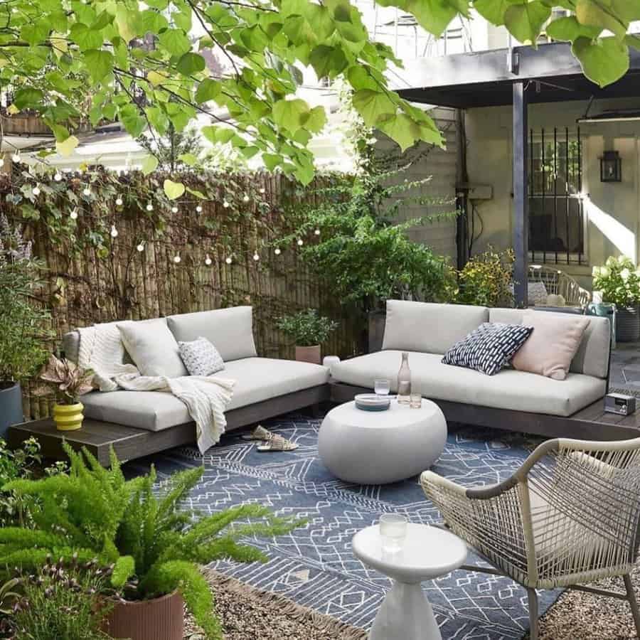 Cozy outdoor patio with gray sofas, a round coffee table, patterned rug, plants, and string lights under leafy trees