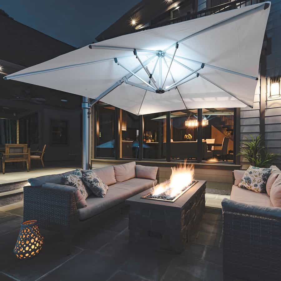 Night patio scene with a lit fire table surrounded by cushioned wicker sofas. A large umbrella is illuminated above, creating a cozy ambiance