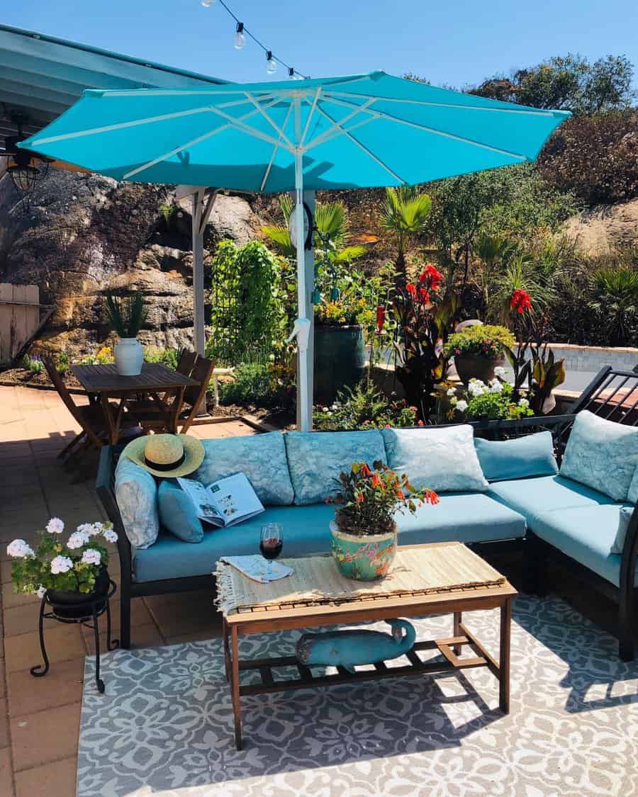 Outdoor patio with blue sectional sofa, table, teal umbrella, and plants. A book and hat rest on the sofa. String lights overhead