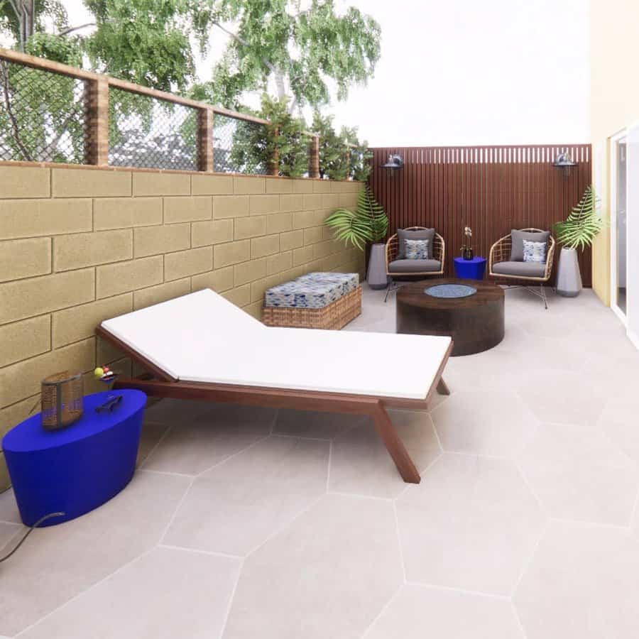 Modern patio with a sun lounger, round fire pit, two chairs, potted plants, and a fence in neutral tones with blue accents