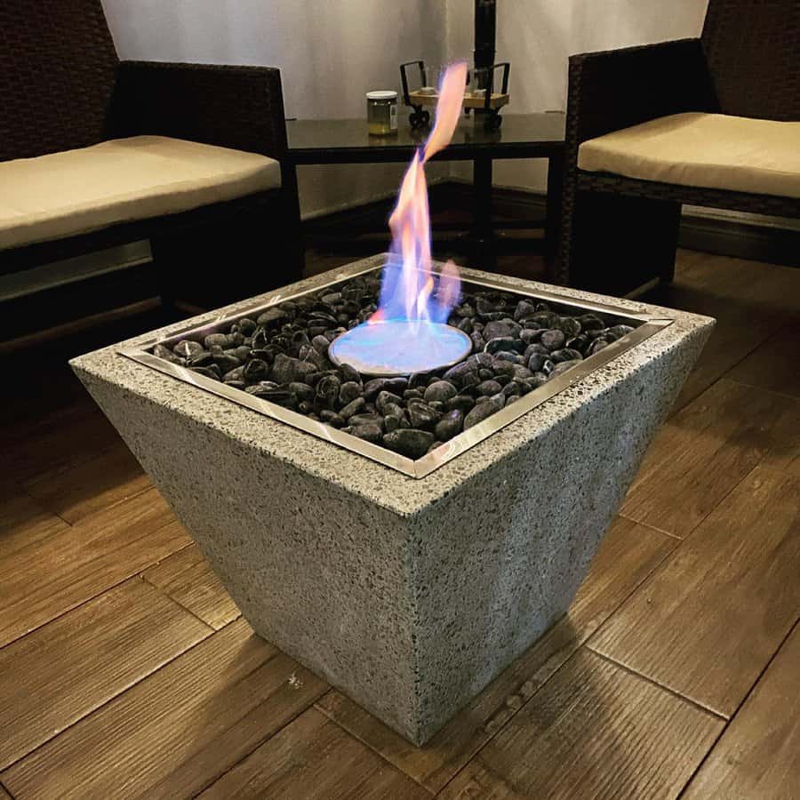 Modern fire pit on a wooden floor, surrounded by chairs, with flames rising above decorative stones