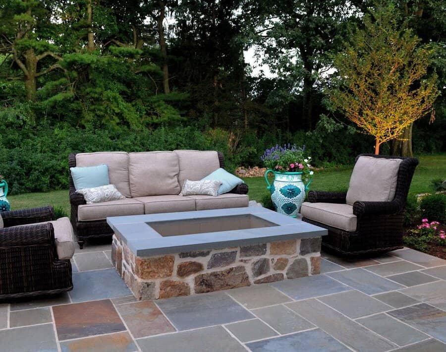 Outdoor patio with wicker furniture, cushions, and a stone fire pit, surrounded by greenery and lit trees in the background