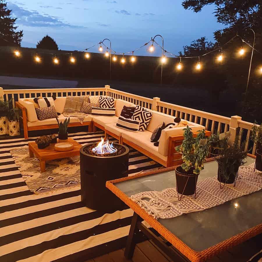 Cozy patio at dusk with string lights, a modern couch, geometric pillows, a fire pit, and a table with potted plants on a striped rug