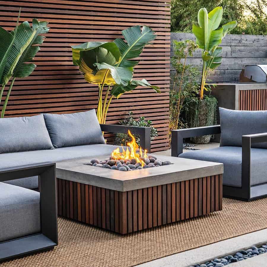 Outdoor patio with modern gray sofas, a rectangular fire pit, wooden slat wall, and tropical plants; cozy seating area