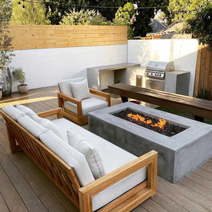 Modern outdoor patio with white cushioned wooden furniture, a concrete fire pit, and a barbecue grill set against a wooden fence backdrop