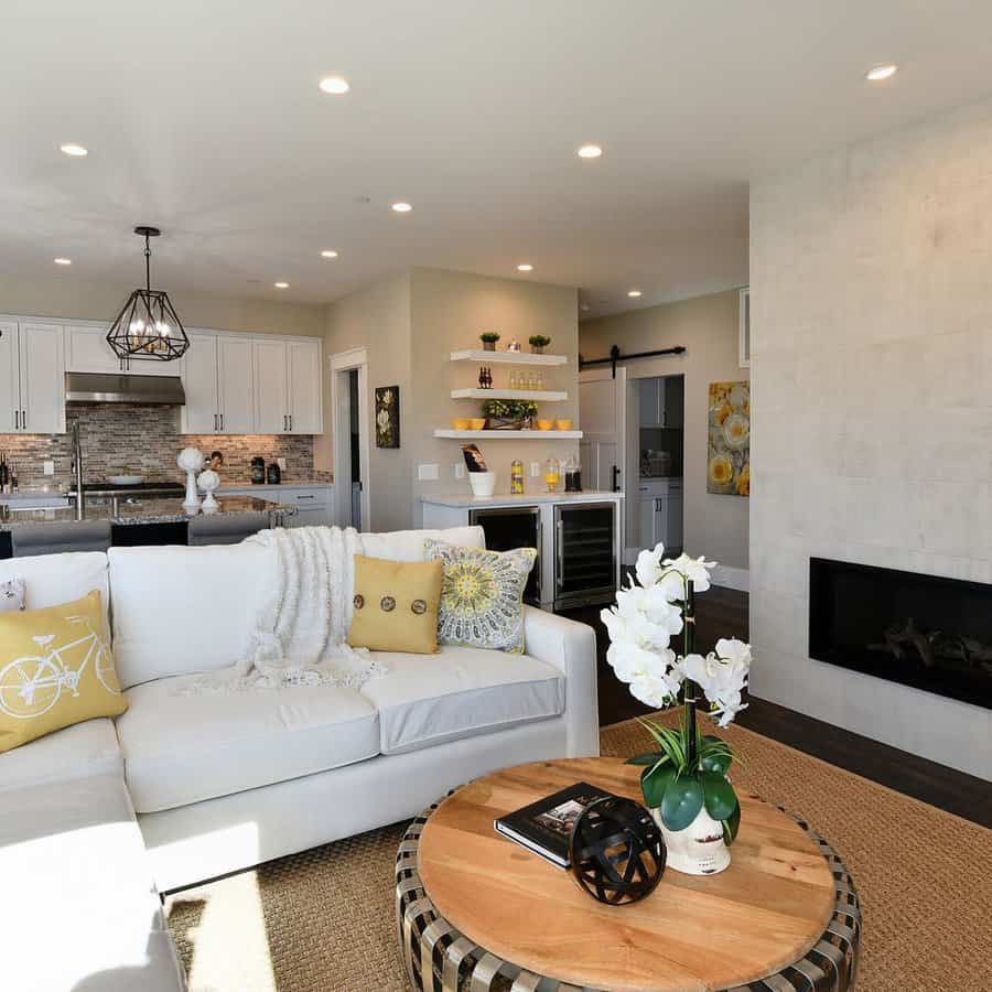 Modern living room with a white sofa, round wooden coffee table, built-in fireplace, and open kitchen featuring yellow accents and orchids