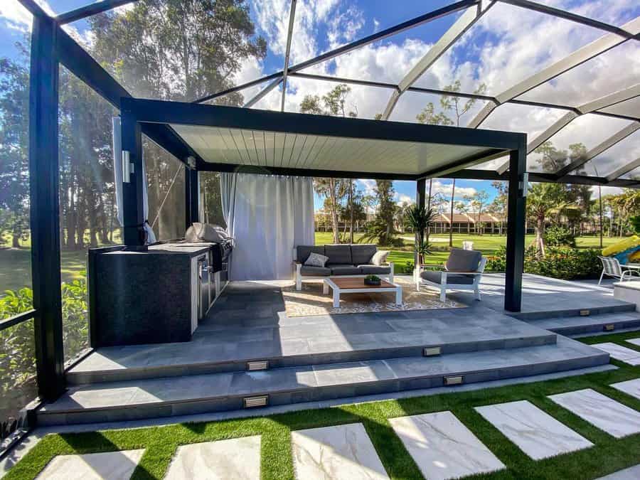 Modern outdoor patio with pergola, couch, and coffee table. Steps lead to grassy area with stone pathway; trees in the background.