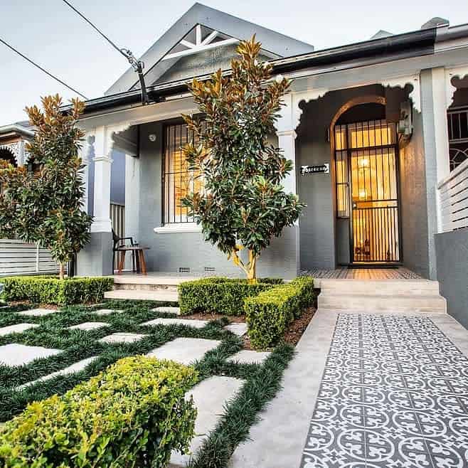 Charming front yard with patterned walkway tiles, manicured hedges, stepping stones with grass inlays, and a porch with decorative arches