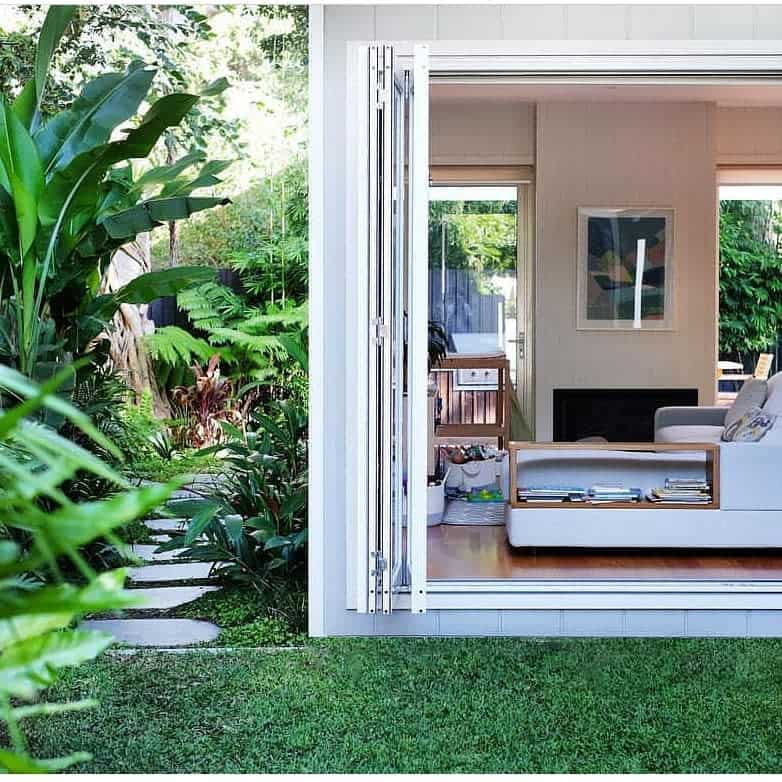 Tropical backyard with lush greenery, stepping stone path, and open living room featuring bi-fold doors for seamless indoor-outdoor flow.