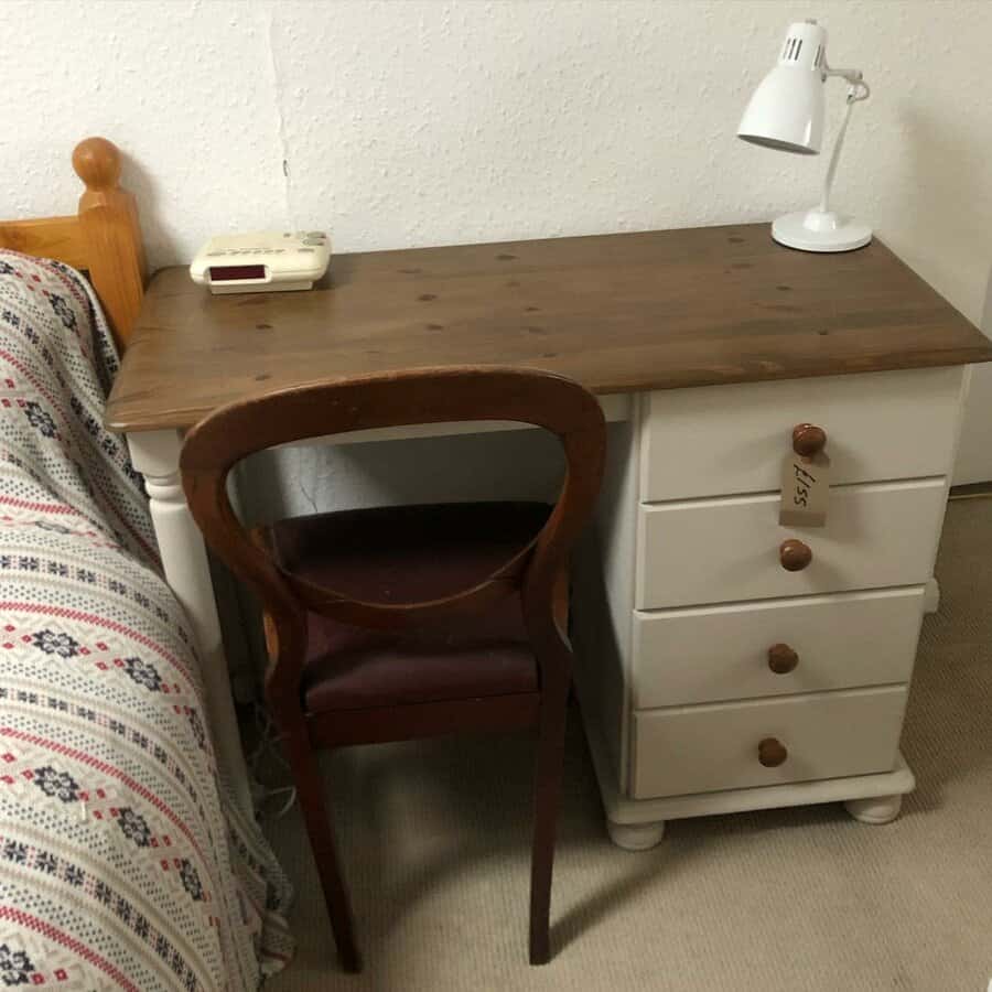 Wooden desk and drawers with a white lamp and phone, alongside a patterned bedspread and wooden chair