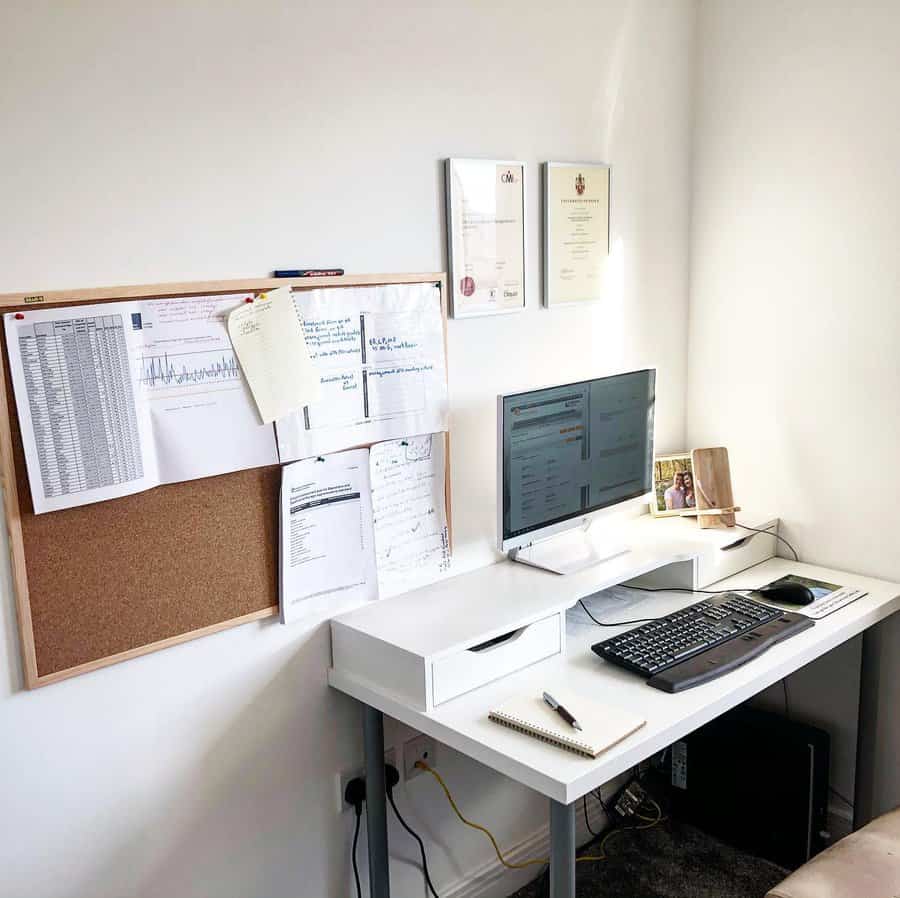 Home office corner with a corkboard, documents, computer on a white desk, and certificates on the wall