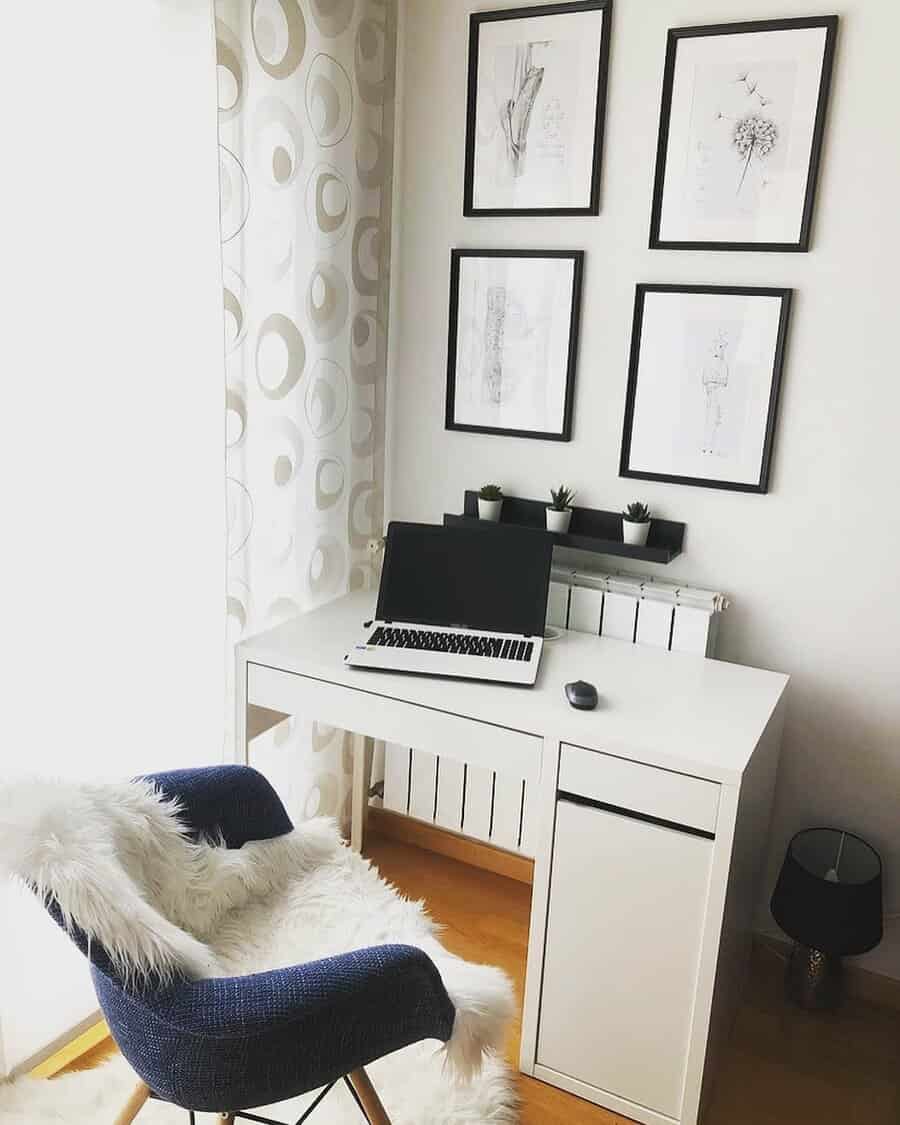 A minimalist home office with a white desk, open laptop, blue chair with a fur throw, and four framed sketches on the wall