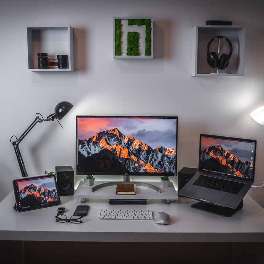 Modern desk setup with monitor, laptop, tablet, keyboard, mouse, and speakers; shelves with decor on white wall background