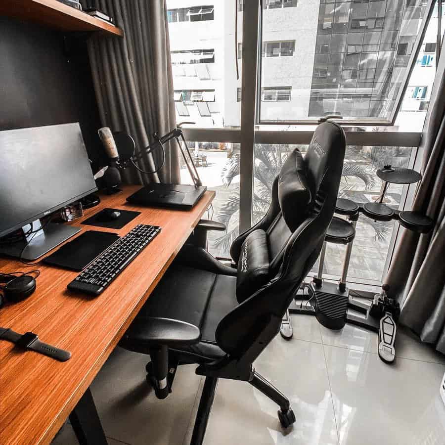 Home office with a wooden desk, computer setup, microphone, and gaming chair; large window with a view of buildings in the background