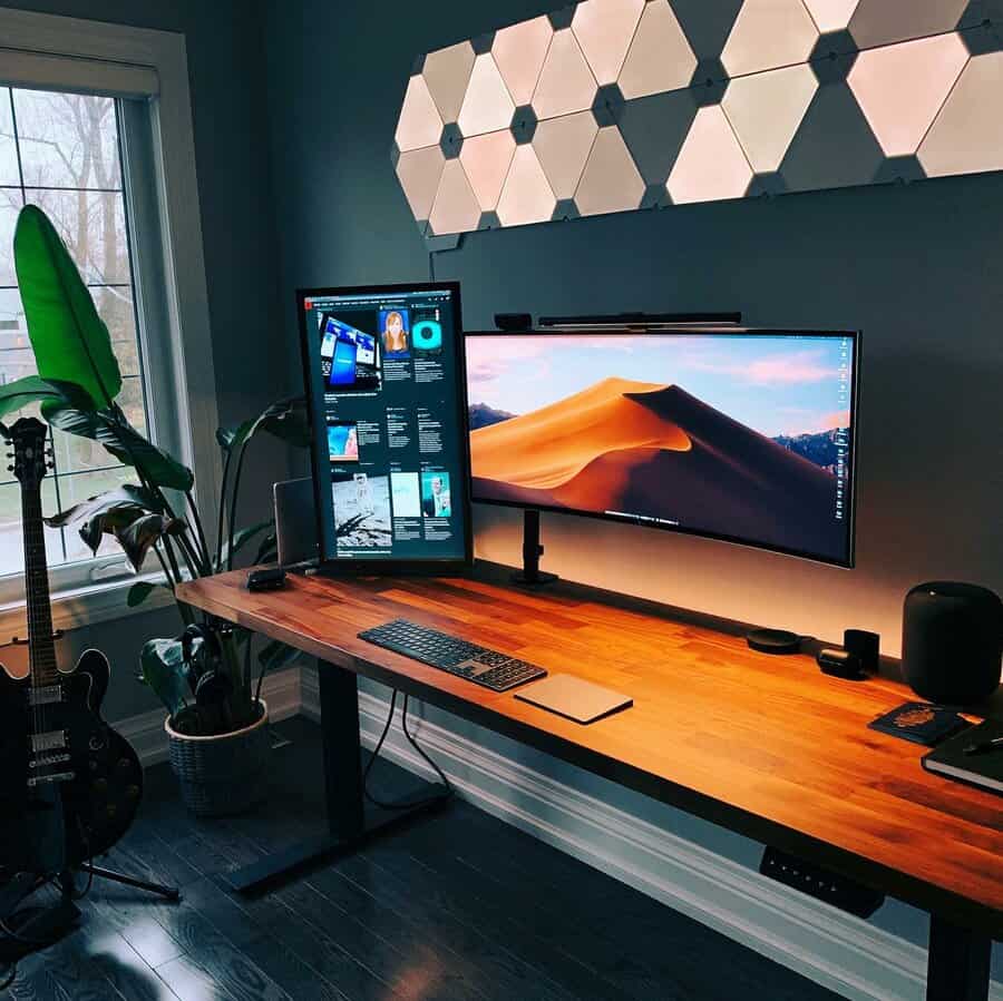 Dual-monitor setup on a wooden desk with keyboard and speakers, guitar nearby, and hexagonal wall lights above