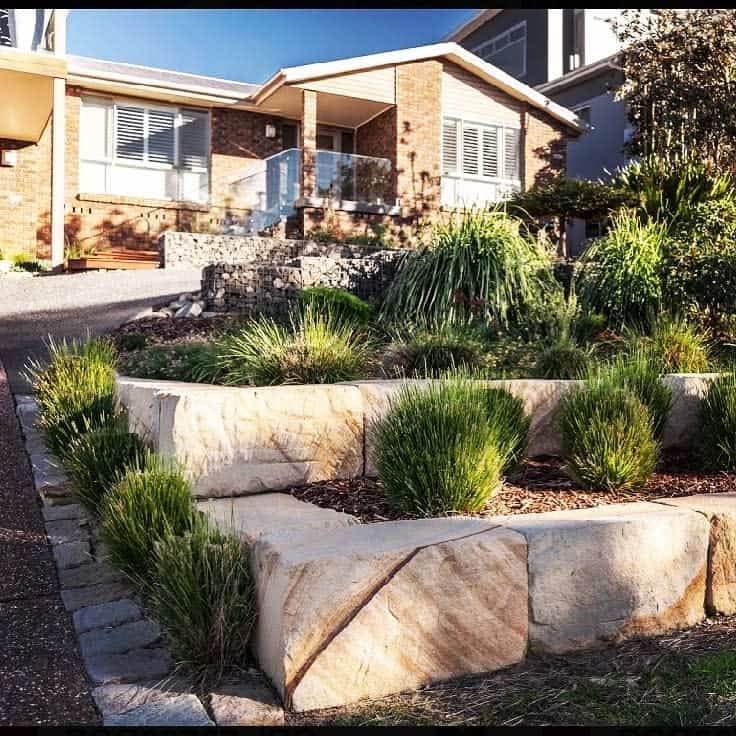 Modern front yard with tiered sandstone retaining walls, drought-tolerant plants, and a stylish brick home in the background