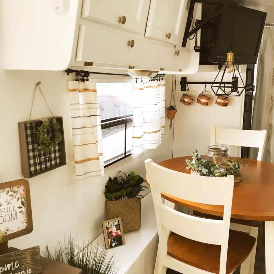 A cozy RV dining area with a wooden table, white chairs, hanging copper mugs, and greenery accents, creating a warm, inviting atmosphere.