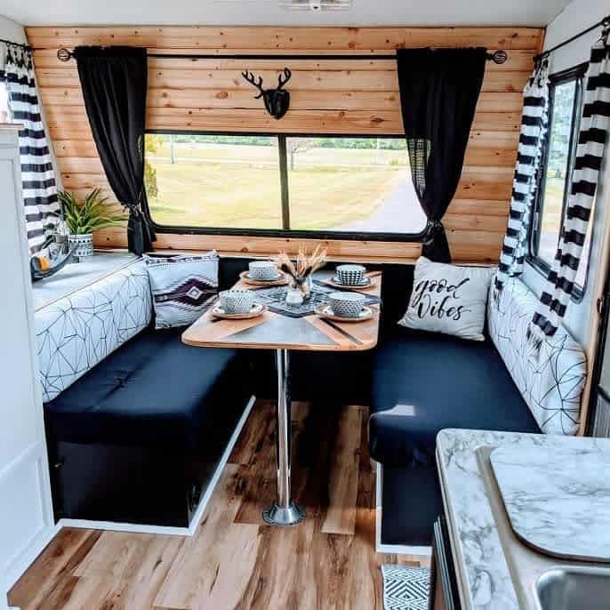 A stylish RV dining area featuring a wooden table, dark seating with geometric pillows, black-and-white curtains, and a deer antler accent, creating a cozy and modern space.