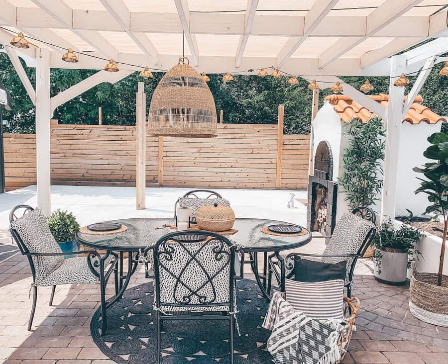 Outdoor patio with a round glass table, patterned chairs, and a woven pendant light, featuring a wooden privacy fence and plants in the background