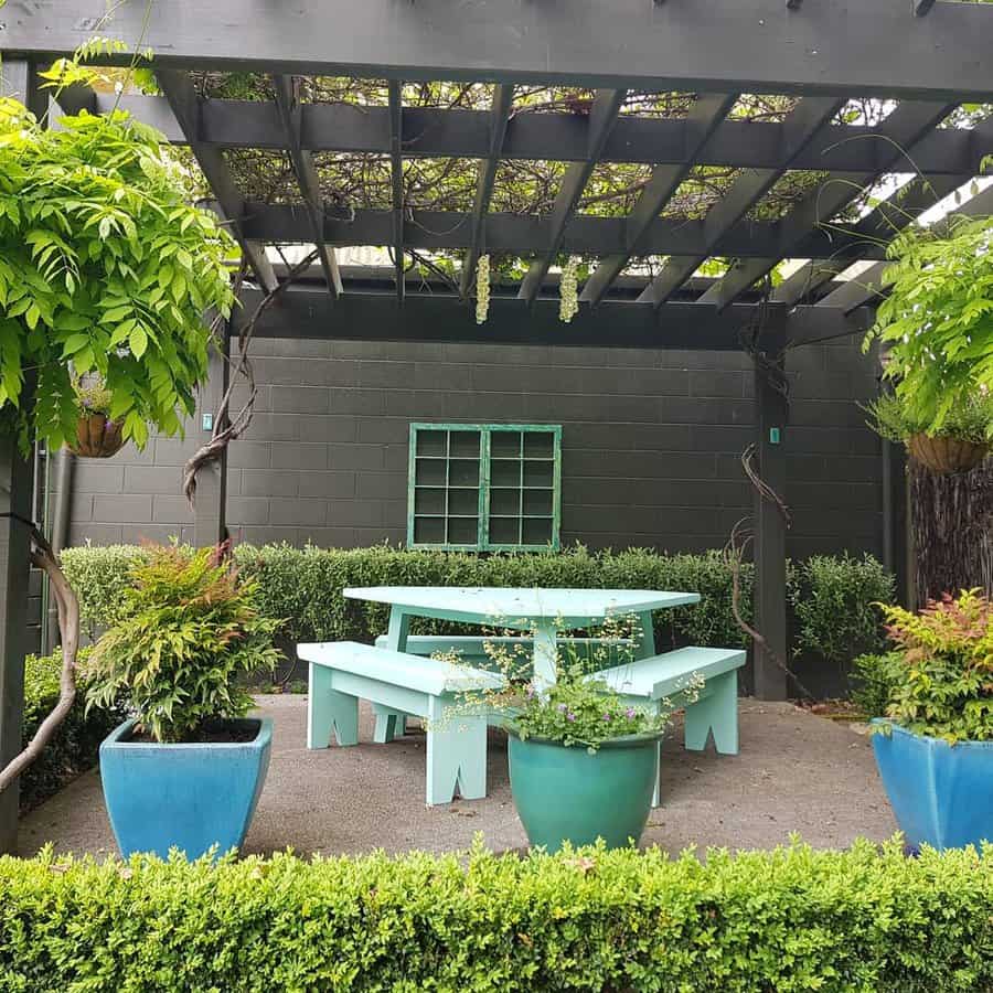 Wooden pergola with greenery overhead, teal picnic table with benches, potted plants, and trimmed hedge in a garden setting