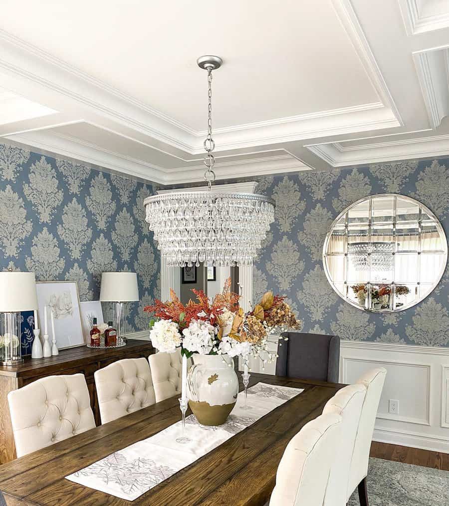 Elegant dining room with a wooden table, white cushioned chairs, floral centerpiece, crystal chandelier, and blue damask wallpaper