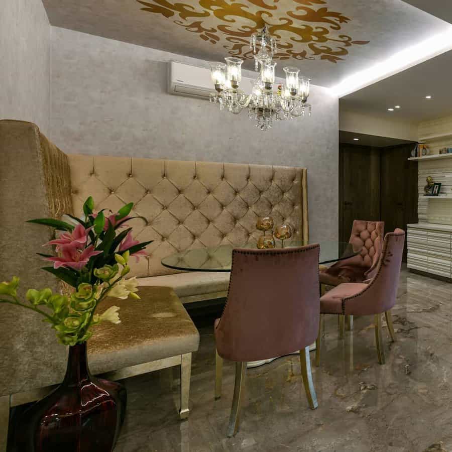 Elegant dining area with tufted bench seating, velvet chairs, glass table, chandelier, and floral arrangement in a vase on marble flooring