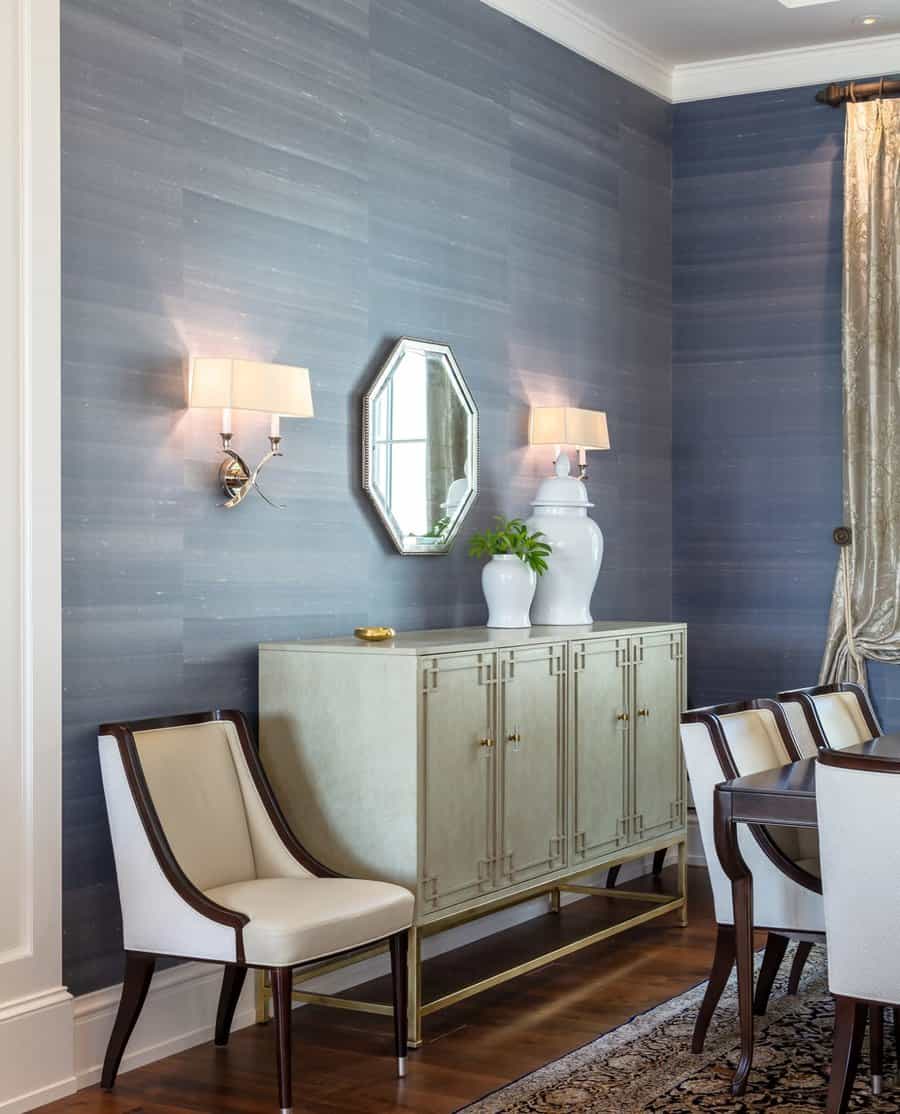 Elegant dining room with blue walls, a hexagonal mirror, sconces, a sideboard with a vase, and an upholstered chair