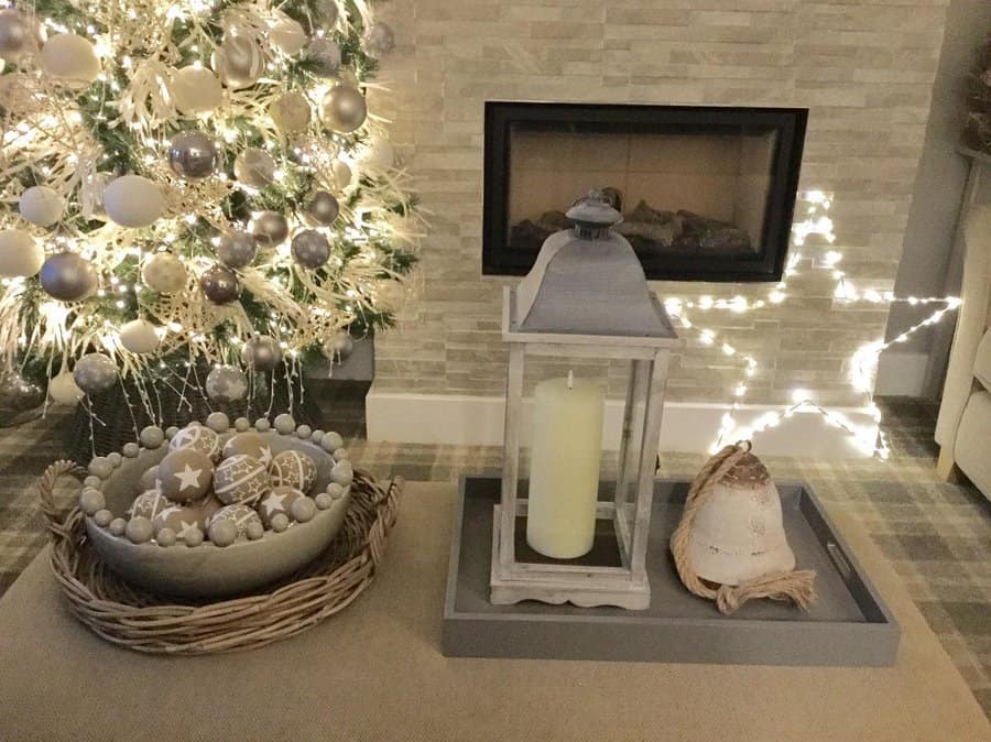 Cozy living room with a Christmas tree, silver ornaments, a lit fireplace, and festive decorations on a tray including a candle and lantern