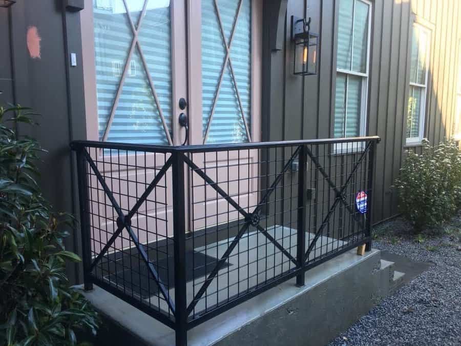 Small porch with black metal railing, glass-paneled pink door, and exterior wall lantern; gravel path and greenery on the side