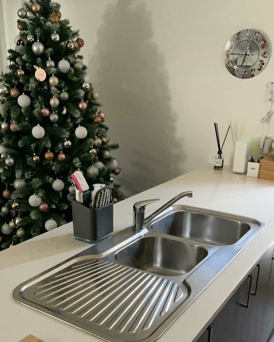 Modern kitchen with a stainless steel double sink and drainboard on a white countertop, with a decorated Christmas tree in the background