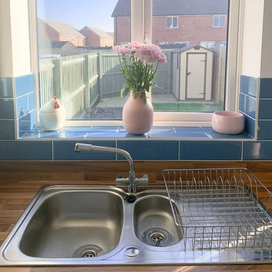 A metal kitchen sink with a drying rack, pink vase with flowers, and jars on a windowsill overlooking a fenced yard and shed