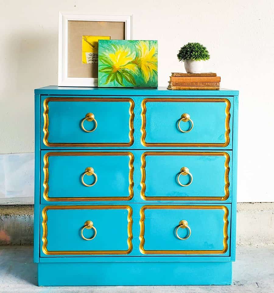 Vibrant turquoise dresser with ornate gold trim and ring pulls, styled with colorful artwork, books, and a small potted plant.