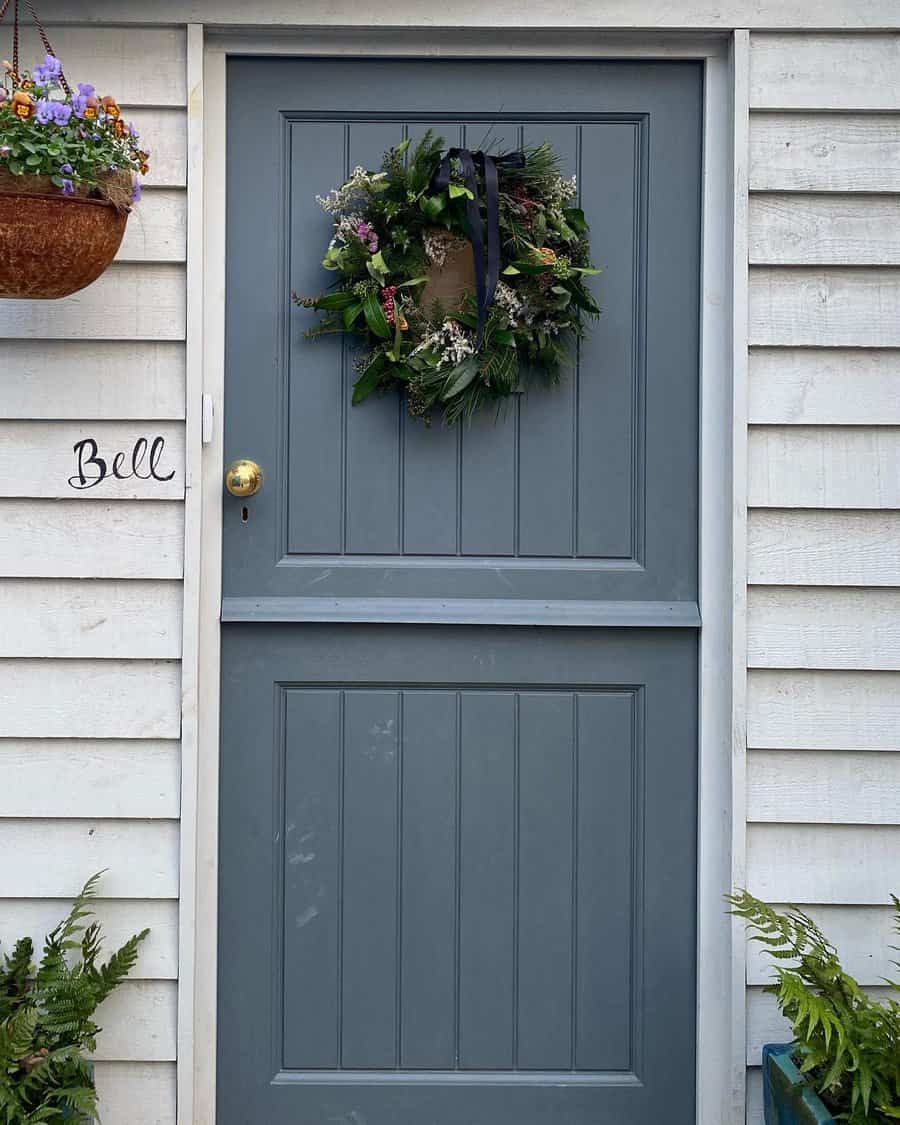 Gray door with a festive wreath and a gold knob on a white paneled wall, hanging basket with flowers to the left, small sign reads "Bell"