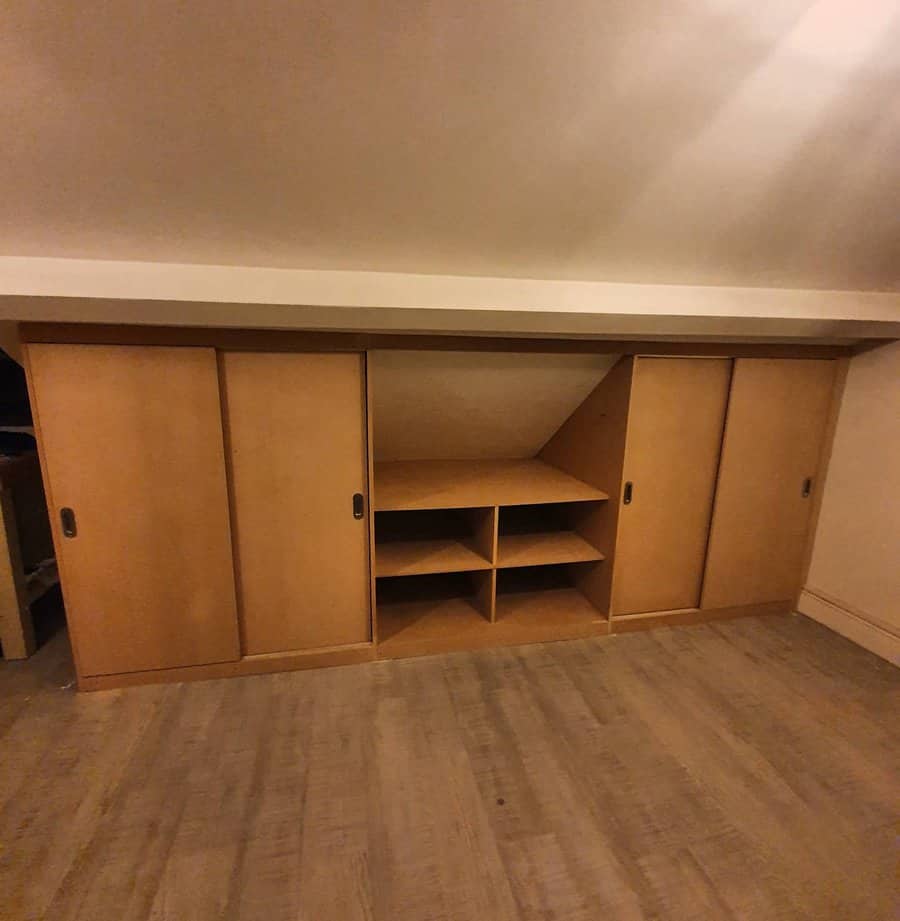 Built-in wooden storage unit with sliding doors and open shelves under a sloped attic ceiling, maximizing space efficiently.