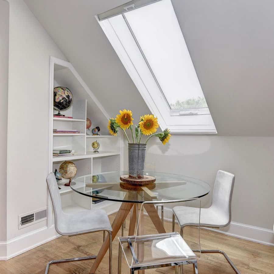 Bright attic room with a glass table, two white chairs, sunflowers in a vase, and a slanted skylight; bookshelf holds globes and decor