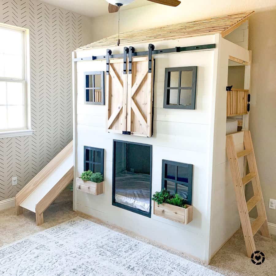 Children's playhouse bed with slide, barn doors, ladder, and plant boxes, situated in a room with a patterned wall and ceiling fan