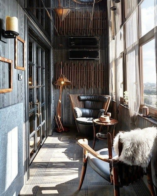 Sophisticated enclosed balcony with dark wood paneling, leather chairs, a cozy fur throw, warm lighting, and floor-to-ceiling windows