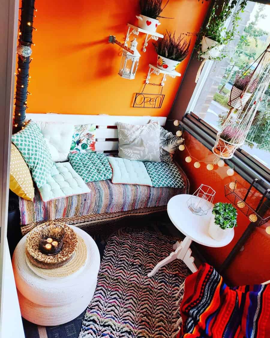 Vibrant enclosed balcony with an orange accent wall, colorful cushions, boho decor, hanging plants, fairy lights, and a cozy seating nook