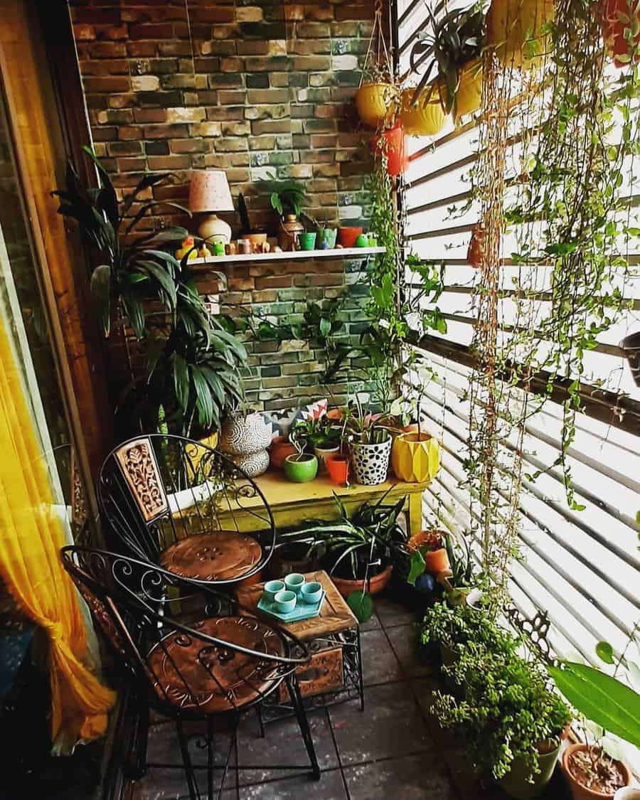 Lush enclosed balcony with a brick accent wall, hanging plants, potted greenery, wrought iron chairs, and a cozy boho seating nook