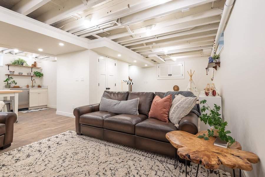 Warm and inviting basement with an open ceiling, cozy leather sofa, natural wood accents, and a functional kitchenette for a stylish retreat.