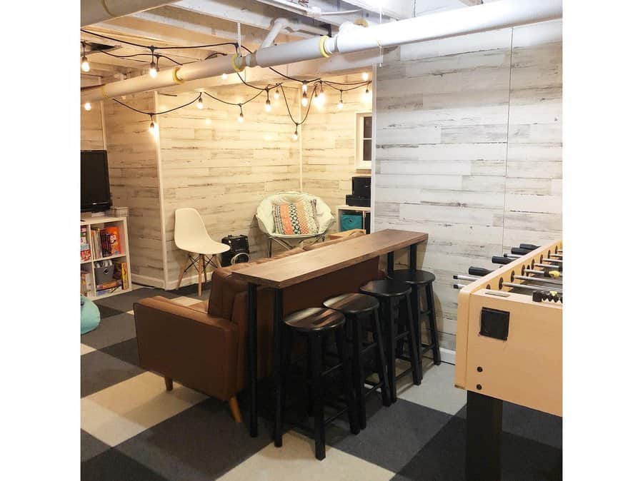 Cozy basement game room with an open ceiling, string lights, wood panel walls, a foosball table, and a lounge area for relaxed entertainment.