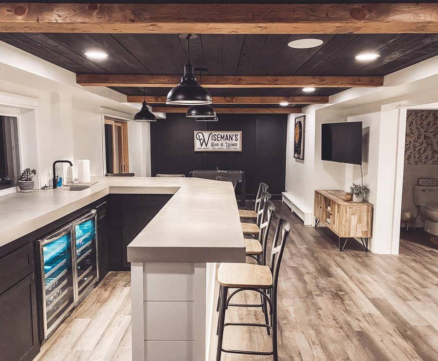 Rustic basement bar with exposed wood beams, concrete countertops, black cabinetry, and industrial lighting for a cozy and stylish lounge space.