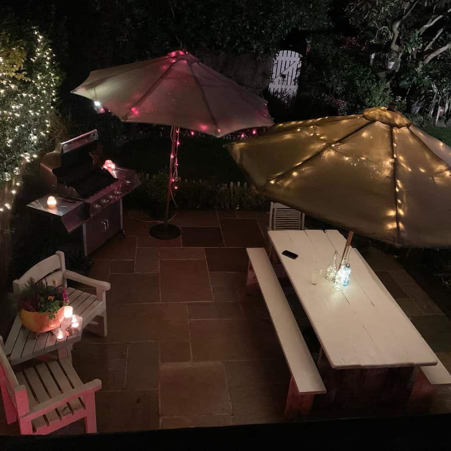 Cozy backyard patio with lit umbrellas, a white picnic table, bench seating, a barbecue grill, and string lights at night