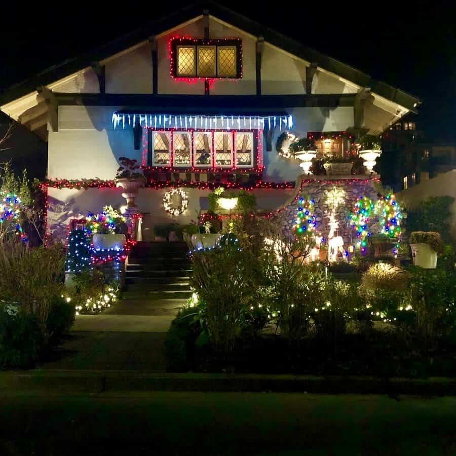 House decorated with colorful Christmas lights and wreaths at night; lit bushes and icicle lights adorn the cozy front porch