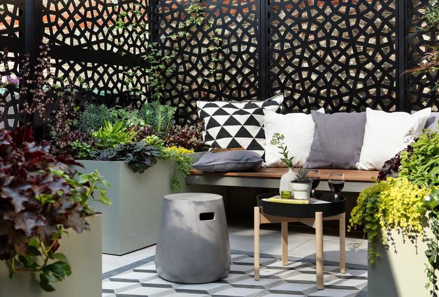 Modern patio with a geometric privacy screen, built-in bench seating, lush planters, monochrome pillows, and a sleek side table