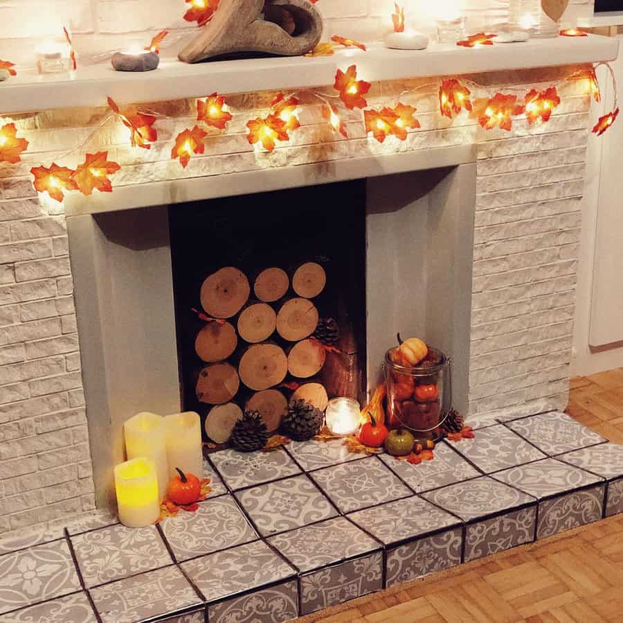 A fireplace decorated with autumn leaves, candles, pumpkins, pinecones, and lights; logs are stacked inside the fireplace